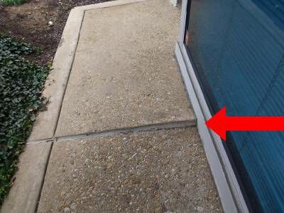 An exposed-aggregate concrete walkway beside a glass wall, with a red arrow at a joint where the slab has dropped
