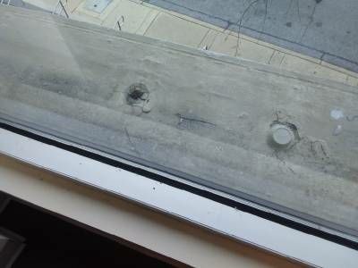 A concrete drainage sill outside an upper-floor window, cracked and broken around a bolt head