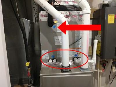 A gas furnace and air handler with white PVC pipes rising from it, a red oval around the openings where the pipes pass through the cabinet and a red arrow pointing to the upper panel