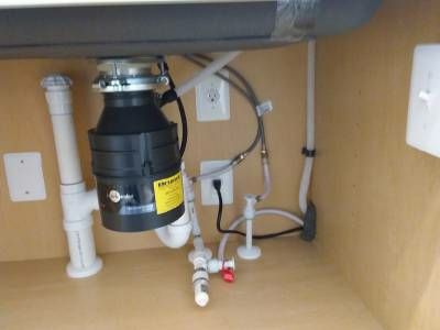 A black garbage disposal mounted beneath a stainless kitchen sink, with white drain pipes, supply lines and wall outlets in the cabinet behind it