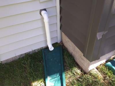 The rear corner of a house where light and dark siding meet above the foundation, with a white sump pump discharge pipe ending over a green splash block on the lawn