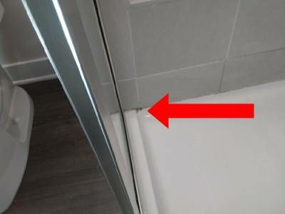 The bottom corner of a glass shower door frame on a white shower base, with a red arrow pointing to the base of the frame