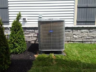 A heat pump condenser on a pad beside a stone-and-siding wall, with a small evergreen planted close to it