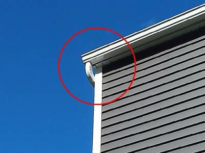 The corner of a house with dark gray siding and white trim against a blue sky, with a red circle around a piece of soffit pulling away at the roof line