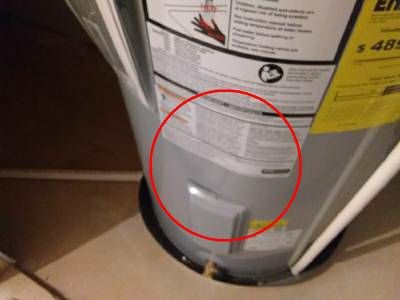 The lower front of a gray electric water heater, with a red circle around a bent section of its jacket above the access panel for the temperature controls